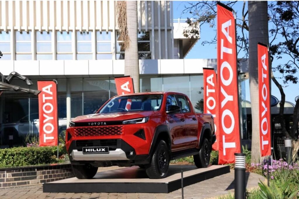A line of newly manufactured Toyota pickup trucks, led by a bright red model with an open driver-side door, moving down a brightly lit factory assembly line.